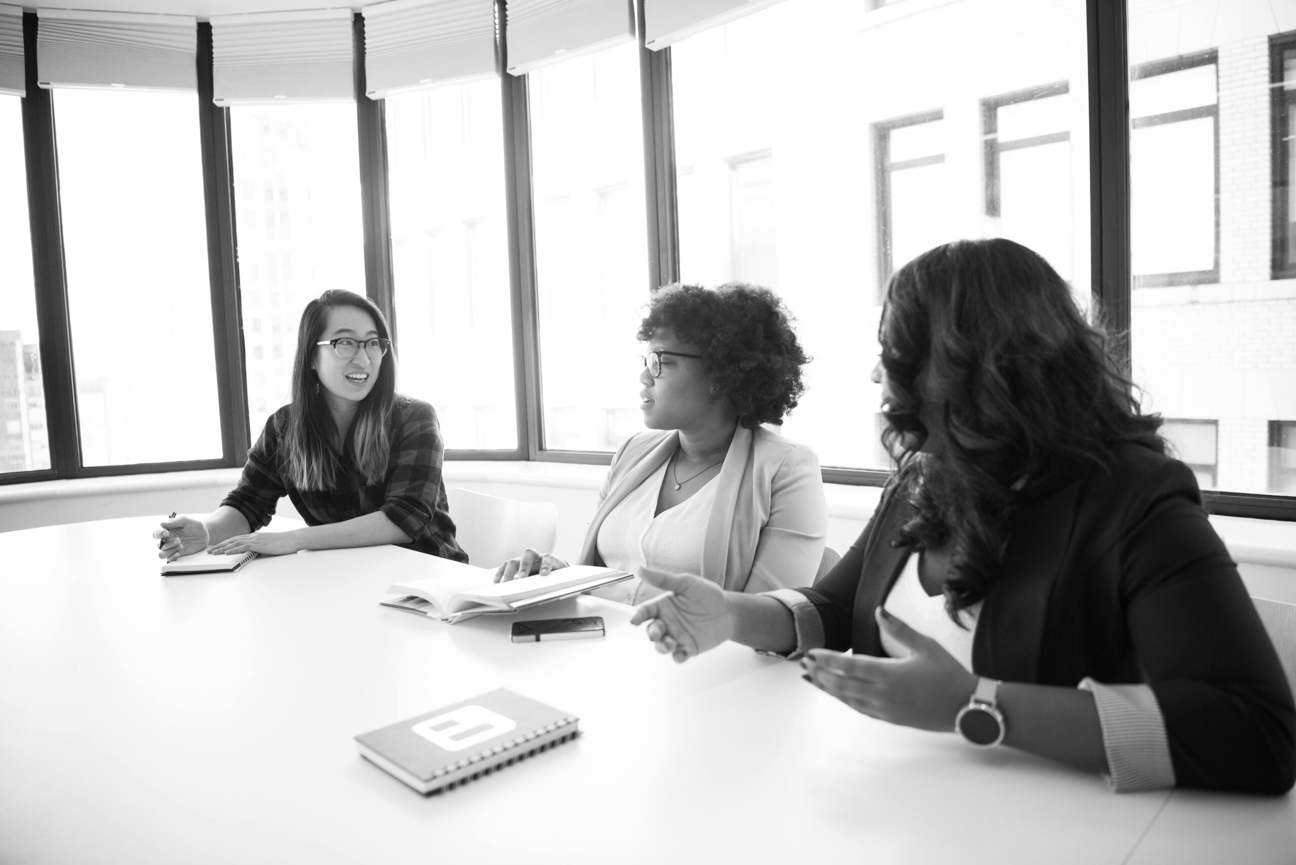 Three women engaged in a business meeting around a table in a modern office setting.